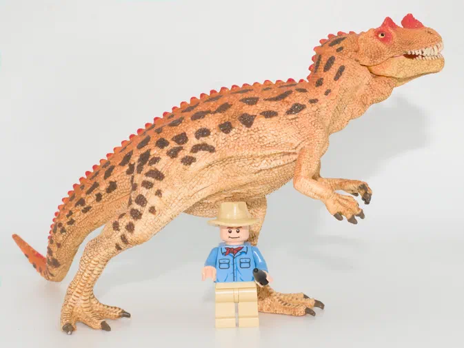 Un grand jouet de dinosaure beige avec des taches brunes sur le dos, se tenant au-dessus d'une petite figurine Lego masculine portant un chapeau et une chemise bleue.