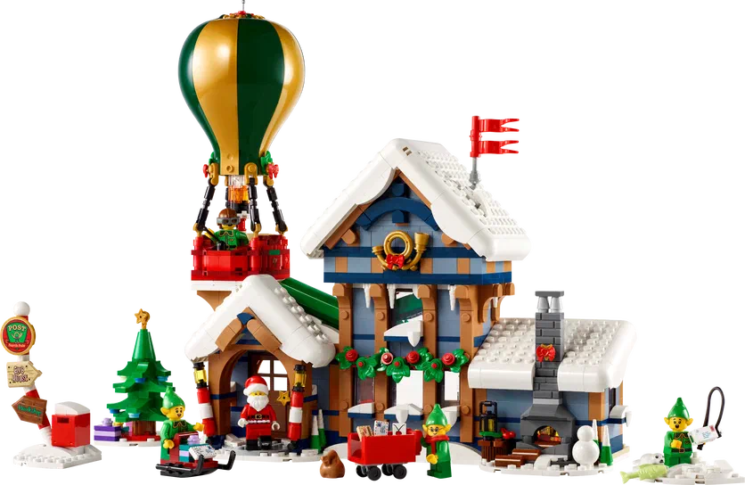 Une maison en bois bleue et blanche avec cheminée décorée pour Noël, Père Noël à l'entrée, des lutins verts autour, un ballon dirigeable vert et doré attaché au toit d'une annexe rouge.