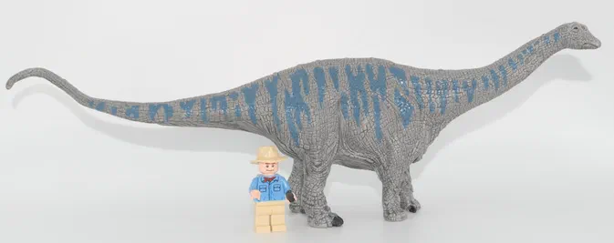 Jouet de dinosaure sauropode gris texturé avec des motifs bleus, photographié sur fond blanc avec une petite figurine homme en costume bleu à côté pour indiquer la taille.