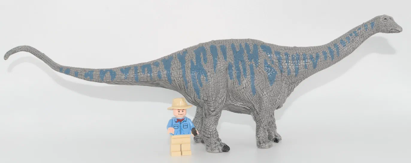 Jouet de dinosaure sauropode gris texturé avec des motifs bleus, photographié sur fond blanc avec une petite figurine homme en costume bleu à côté pour indiquer la taille.