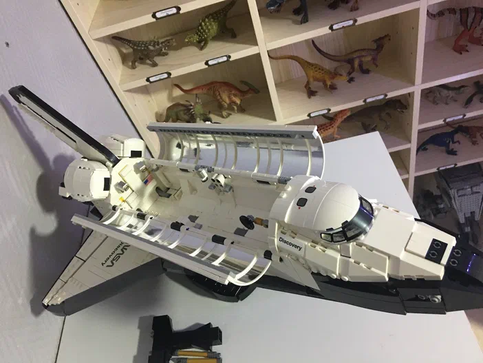 Un modèle de fusée spatiale LEGO blanc et noir nommé Discovery est posé sur une surface blanche, devant des étagères floues remplies d'objets.