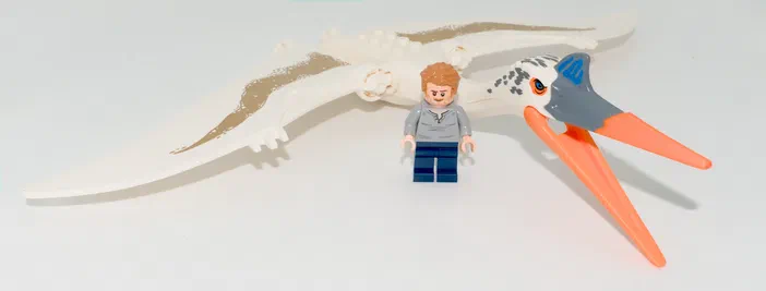Un minifigurine LEGO avec des cheveux roux et un t-shirt gris se tient devant une créature volante blanche à bec orange, issue du set Jurassic World.