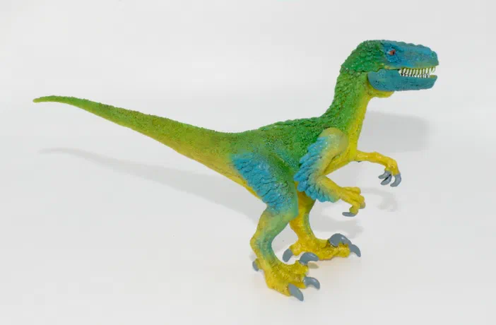 Un jouet de dinosaure bipède aux couleurs vertes et bleues sur fond blanc.