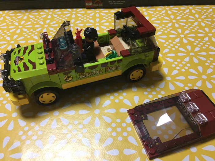 Véhicule vert aux motifs zébrés du jeu Jurassic Park avec une figurine au volant tenant un pistolet rouge, son toit transparent reposant à côté sur le fond jaune floral.