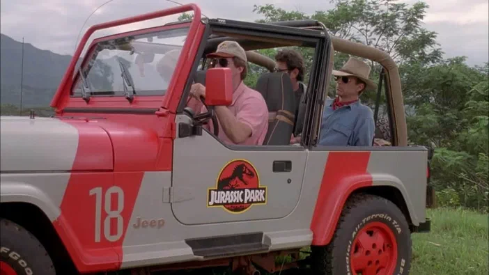 Une Jeep rouge et grise affichant le logo Jurassic Park sur sa porte, occupée par plusieurs passagers au volant d'un véhicule dans un environnement naturel.