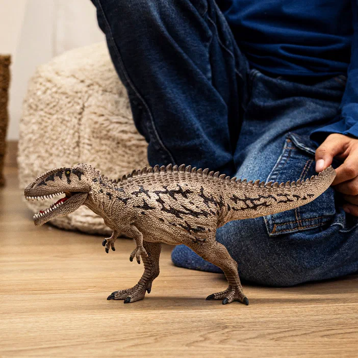 Une main retient par la queue un jouet de dinosaure à deux pattes couleur beige avec des motifs sombres et une crête dorsale pointue, posé sur du parquet clair devant un coussin.