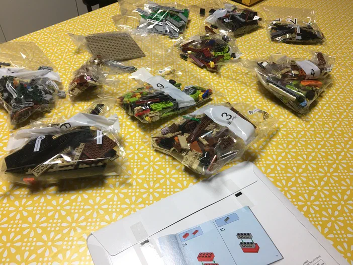 Plusieurs sachets transparents remplis de briques LEGO numérotés sont disposés sur un tapis jaune à motifs floraux blancs, accompagnés d'une page d'instructions ouverte en bas à droite.