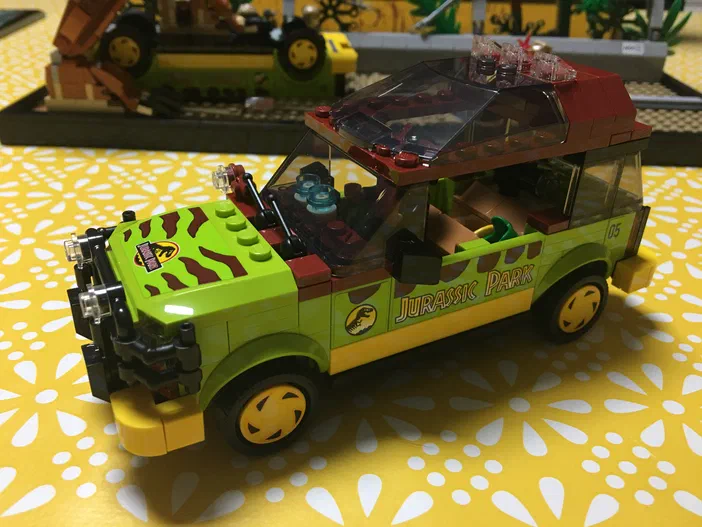 Un modèle de voiture verte LEGO Technic portant l'inscription "JURASSIC PARK" et des rayures marron, posé sur une surface jaune à motifs blancs.