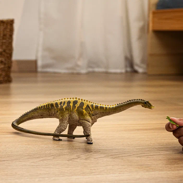 Un modèle jouet d'un dinosaure diplodocus à long cou est posé sur du parquet en bois, face à une main tenant quelques feuilles vertes.