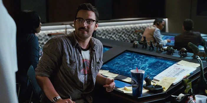 Un homme avec une moustache et des lunettes est assis devant un grand écran bleu affichant une carte ou un plan, entouré de nombreuses petites figurines de dinosaures sur le bord d'un bureau.