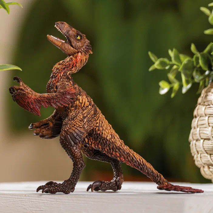 Un jouet représentant un Dakotaraptor à plumes brunes et rouges, debout sur deux pattes avec les bras levés et la bouche ouverte, posé sur une surface blanche devant un fond de verdure.