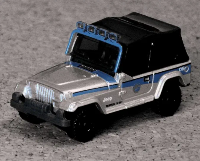 Voiture miniature Matchbox Jeep Wrangler de couleur argentée avec des détails et rayures bleus, photographiée sur un fond gris.