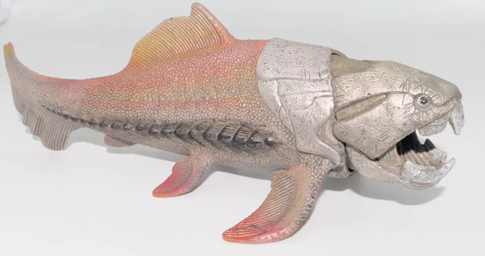 Figurine d'un dinosaure à tête grise avec dents proéminentes et corps rougeâtre écailleux sur fond blanc.