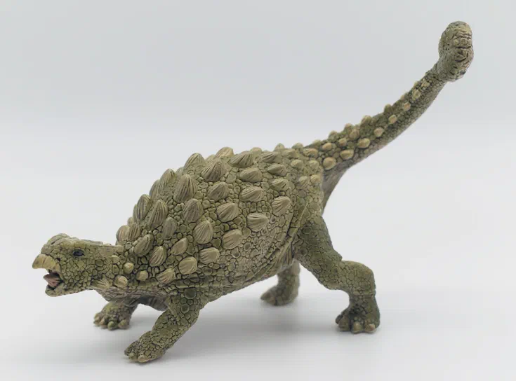 Un jouet de dinosaure vert aux écailles et épines dorsales, avec la queue relevée, posé sur un fond blanc uni.