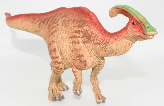 Une figurine d'un Parasaurolophus Schleich debout sur fond blanc, avec une crête verte et un corps beige tacheté.