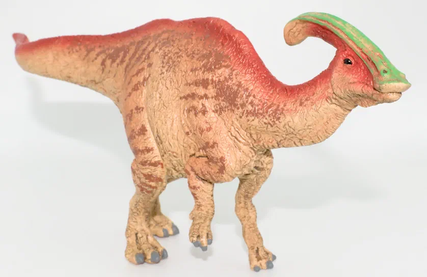 Une figurine d'un Parasaurolophus Schleich debout sur fond blanc, avec une crête verte et un corps beige tacheté.