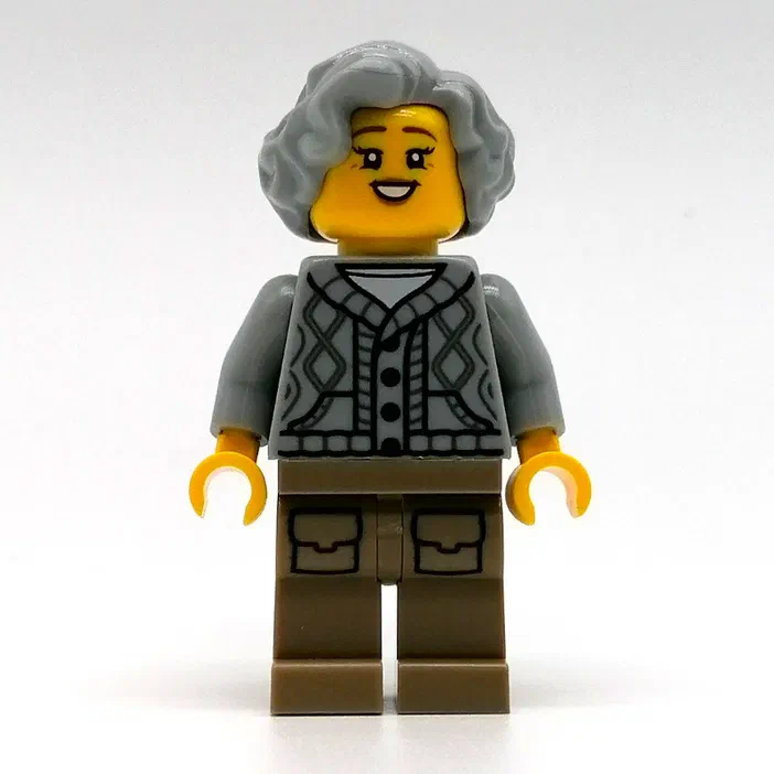 Une minifigurine LEGO d’un personnage féminin aux cheveux gris bouclés portant un gilet tricoté noir sur une chemise grise et un pantalon marron à poches dessinées, posée sur fond blanc.