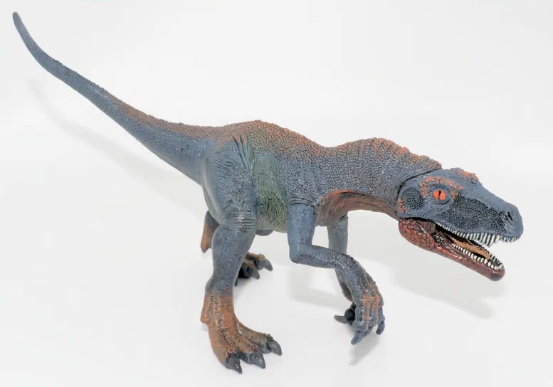 Figurine de dinosaure à deux pattes avec un corps bleu et des accents orange sur le museau et les membres, posée sur fond blanc.