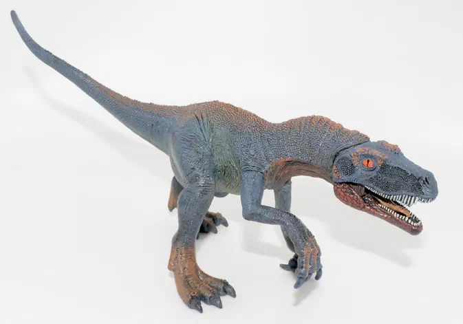 Figurine de dinosaure à deux pattes avec un corps bleu et des accents orange sur le museau et les membres, posée sur fond blanc.