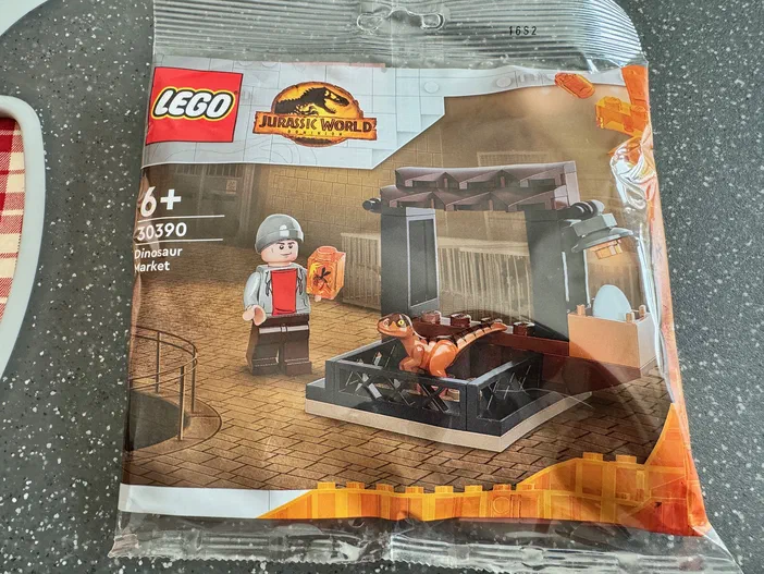 Sac de construction LEGO Jurassic World Dinosaur Market avec un dinosaure orange dans une cage et un minifigurine tenant un conteneur.