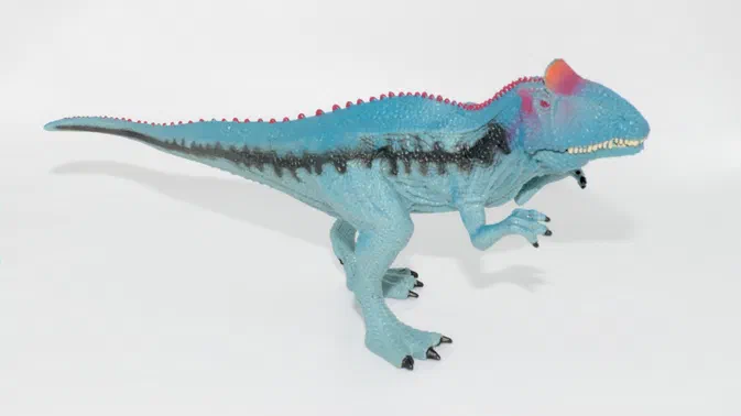 Figurine de dinosaure bleu avec des crêtes rouges et une bande noire sur le dos, posée sur fond blanc.