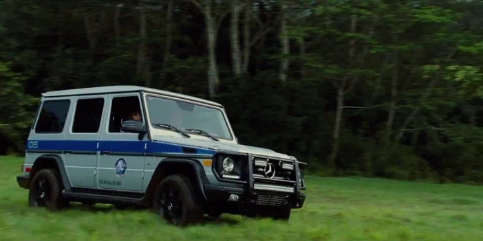 Un véhicule de type G-Wagon bleu et blanc avec des inscriptions officielles roule sur une pelouse verte devant un fond forestier.