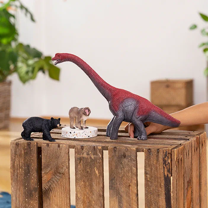 Une main tient un jouet de dinosaure, un brachiosaure rouge et gris, sur une caisse en bois avec d'autres figurines.