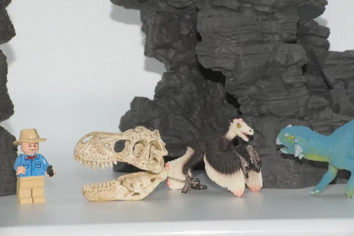Une figurine de minifigurin LEGO habillée en explorateur se tient à côté d'un crâne de dinosaure, une créature ailée et un autre dinosaure bleu devant des rochers gris.