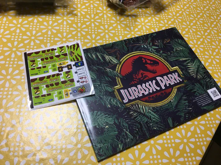 Un ensemble de stickers et d'affiches du jeu LEGO Jurassic World, avec le logo T-Rex sur fond tropical.