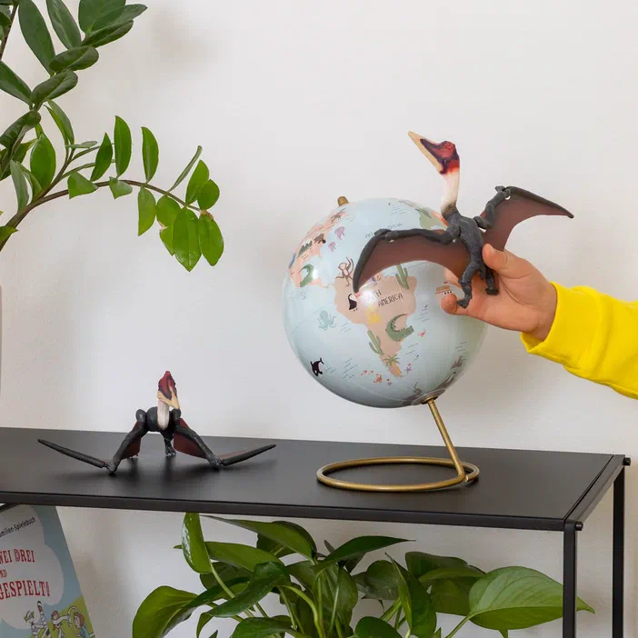 Une main tient un modèle de Quetzalcoatlus devant une boule terrestre, tandis qu'un autre modèle est posé sur l'étagère.