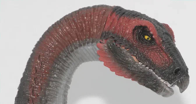 Tête et cou d'un dinosaure jouet aux couleurs rouge, noir et gris sur fond blanc.