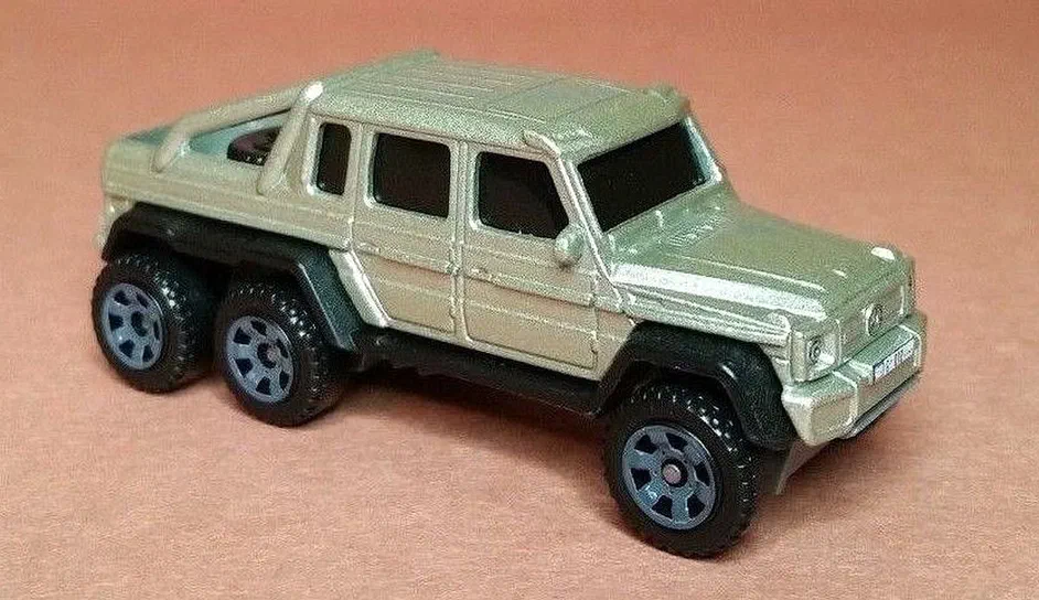 Petite voiture Matchbox Mercedes-Benz G63 AMG 6x6 de la série Jurassic World en beige métallique sur fond rose.