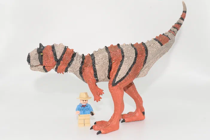 Un jouet de dinosaure à rayures orange et noir avec un petit personnage Lego debout devant lui sur fond blanc.