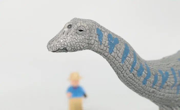 Un jouet d'un Apatosaurus gris avec des marques bleues sur le cou, en gros plan. Un petit personnage flou se trouve derrière.