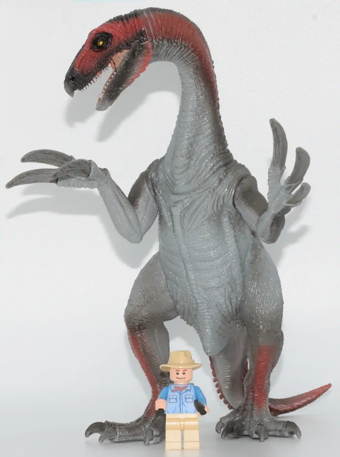 Un dinosaure gris à tête rouge, avec des griffes et une queue visibles, se tient debout sur un fond blanc. Un petit personnage en costume bleu est placé devant lui.