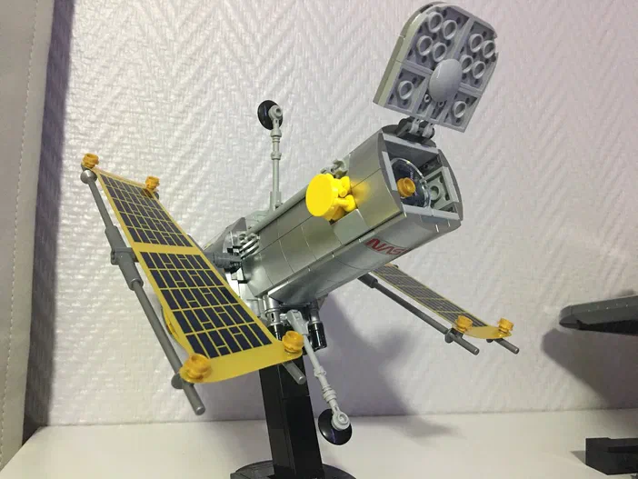 Un modèle de satellite en LEGO avec des panneaux solaires jaunes et une antenne grise, monté sur un support noir.