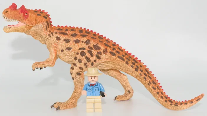 Un dinosaure jouet orange à taches brunes avec une crête rouge et un petit personnage LEGO en costume bleu devant lui sur fond blanc.