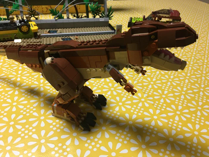 Un dinosaure T-Rex en briques LEGO de couleur marron, posé sur un fond jaune à motifs floraux blancs.