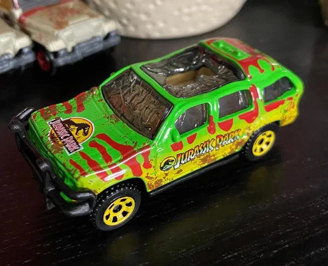 Petite voiture Matchbox verte et jaune avec des rayures rouges représentant un Ford Explorer de Jurassic Park sur une surface sombre.