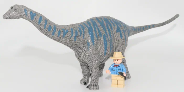 Un dinosaure gris à motifs bleus avec un petit personnage en costume bleu et chapeau beige devant lui.