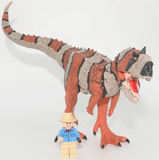 Un jouet de dinosaure T-Rex avec des rayures orange et grises, posé sur un fond blanc. Un petit personnage en plastique portant un chapeau beige se trouve devant le dinosaure.