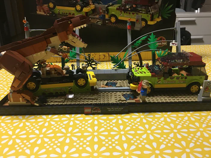 Une scène de jeu LEGO Jurassic World montrant un T-Rex attaquant une jeep verte avec des personnages et des palmiers.