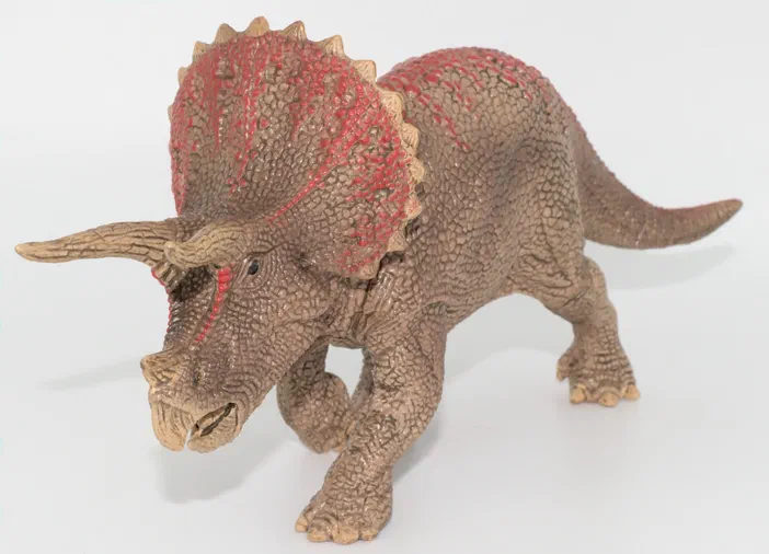 Un jouet de dinosaure triceratops en plastique avec une texture écailleuse et des accents rouges sur le crâne, posé sur fond blanc.