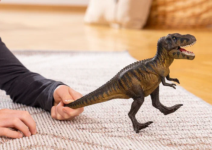 Une main tient un jouet de dinosaure Tarbosaurus sur une tapisserie.