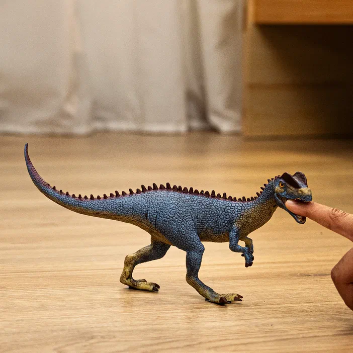 Un jouet de dinosaure Dilophosaurus bleu avec des crêtes dorsales marron est tenu par un doigt sur une surface en bois.
