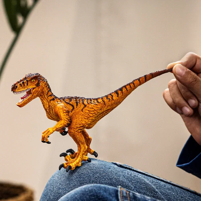 Une main tient un jouet de dinosaure représentant un vélociraptor orange et noir.