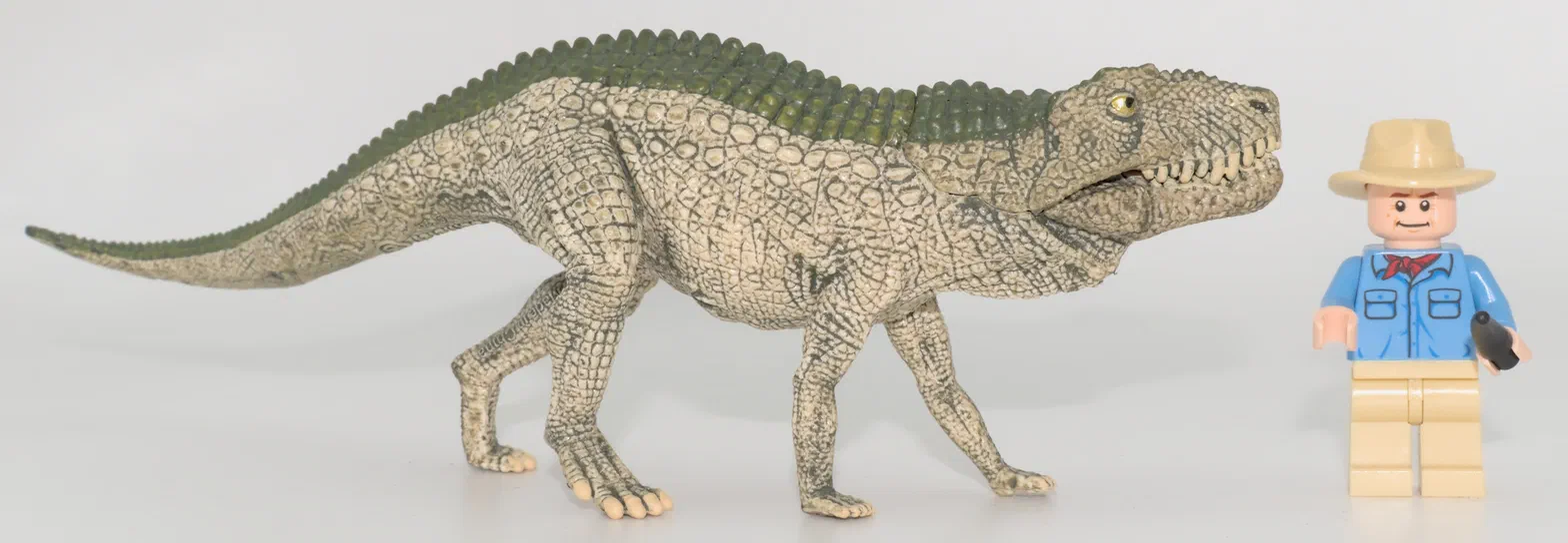 Un dinosaure à quatre pattes de couleur beige et vert est photographié sur un fond blanc avec une figurine Lego en costume bleu.