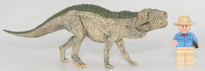 Un dinosaure à quatre pattes de couleur beige et vert est photographié sur un fond blanc avec une figurine Lego en costume bleu.