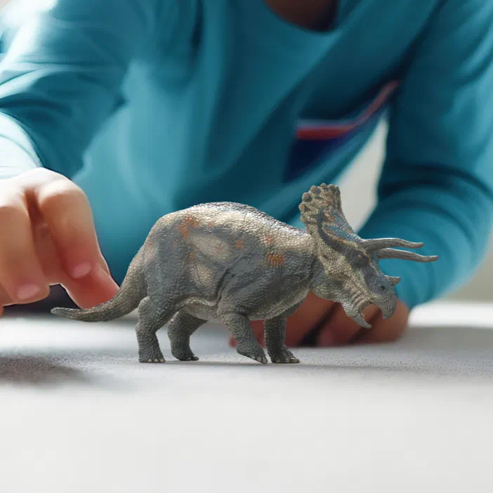 Un jouet Tricératops gris avec des marques orange, posé sur une surface blanche. Une main humaine est visible à gauche du dinosaure.