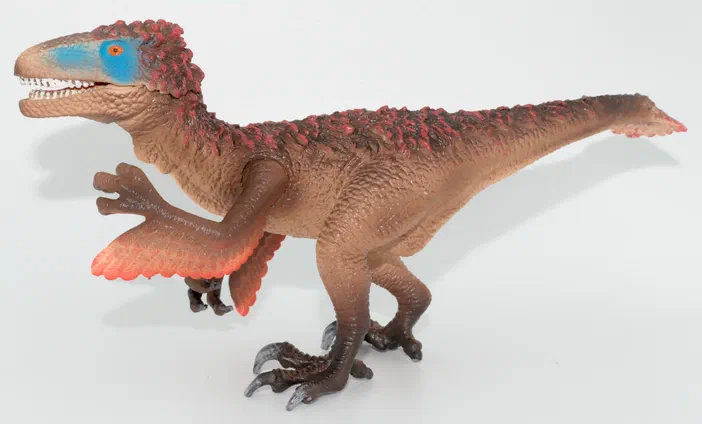 Un jouet de dinosaure bipède à la peau marron avec des crêtes rouges et une tache bleue sur le visage.